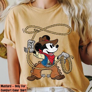 May include: Mustard-colored t-shirt featuring a cartoon cowboy Mickey Mouse with a rope. Mickey is wearing a brown hat, vest, and pants, a red bandana, and gloves. The shirt's text reads "Mustard - Only For Comfort Color Shirt."