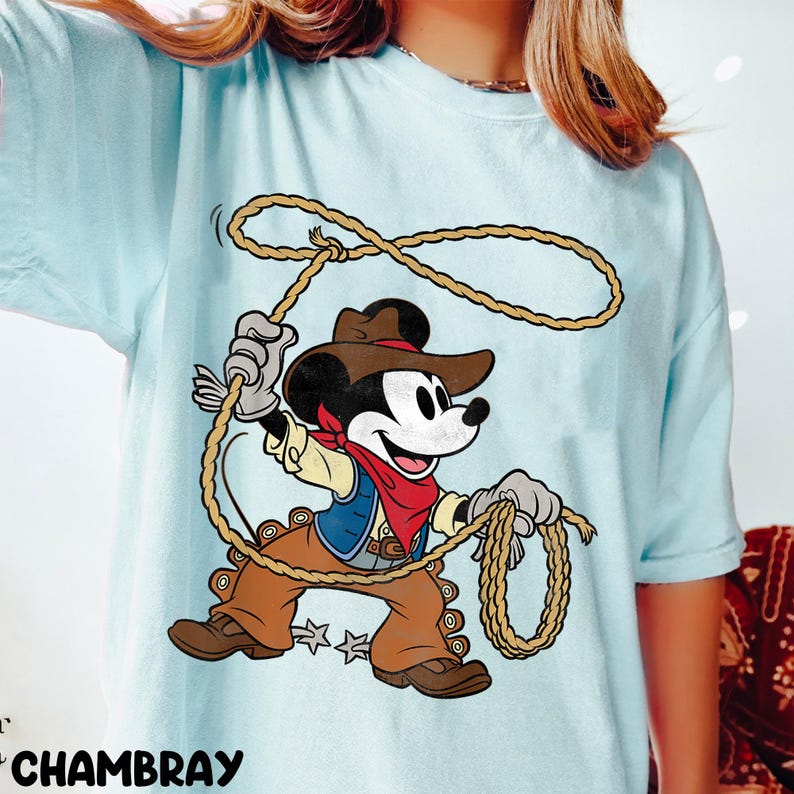 May include: Light blue t-shirt featuring a cartoon Mickey Mouse dressed as a cowboy, holding a lasso. Mickey wears a brown hat, vest, and pants, with a red bandana. The shirt has the word "CHAMBRAY" printed at the bottom.