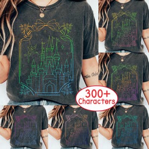 Puede incluir: Camiseta gris oscuro con un diseño de castillo colorido, un lazo y detalles florales. El castillo está delineado en un degradado de colores, con una silueta de hada. La camiseta tiene un aspecto vintage.