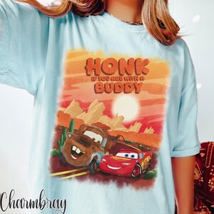 May include: Light blue t-shirt featuring a graphic of two cartoon cars, one brown and one red, with the text "HONK IF YOU ARE WITH A BUDDY" in orange. The design is set against a desert landscape with a sunset.