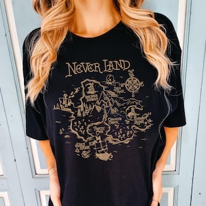 May include: Black t-shirt with a tan map of Neverland. The map design includes the text "Never Land", "Lost Boys Hideout", "Skull Rock", "Mermaid Lagoon", and "Pirate Cove". The shirt has a crew neck.