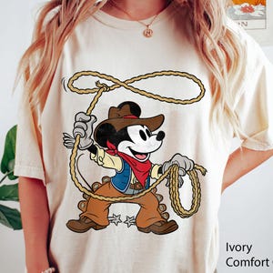 May include: Ivory Comfort Colors t-shirt featuring a cartoon Mickey Mouse dressed as a cowboy, holding a lasso. Mickey wears a brown hat, vest, and pants, with a red bandana. The shirt has the words "Ivory Comfort" printed on the bottom right.