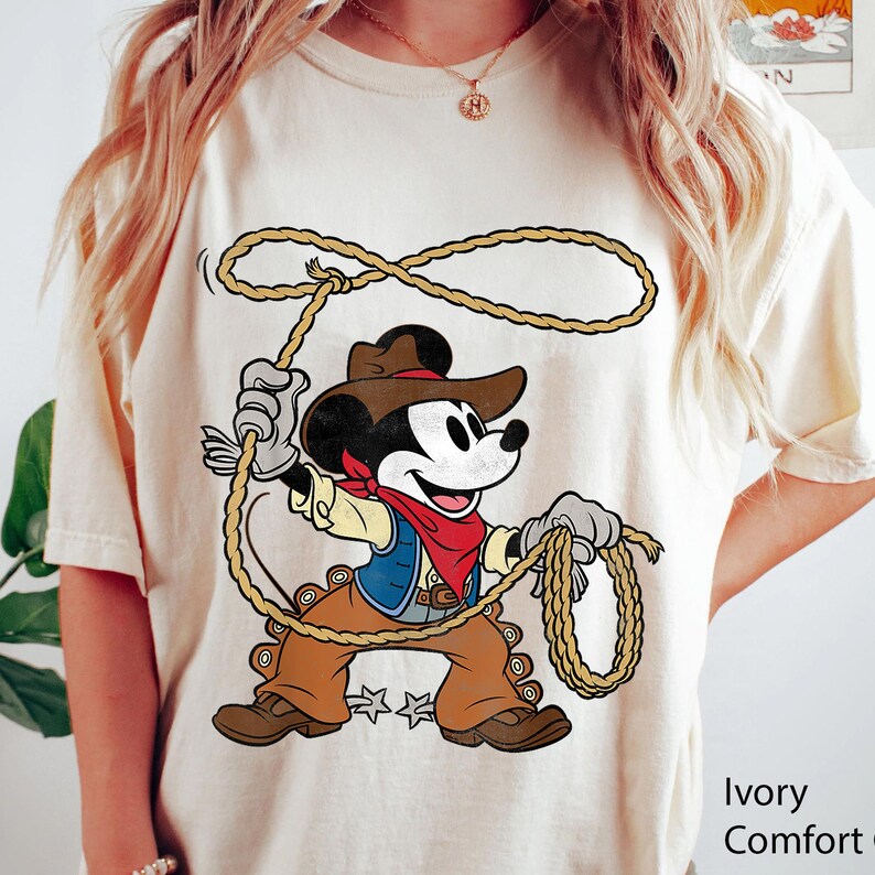 May include: Ivory Comfort Colors t-shirt featuring a cartoon Mickey Mouse dressed as a cowboy, holding a lasso. Mickey wears a brown hat, vest, and pants, with a red bandana. The shirt has the words "Ivory Comfort" printed on the bottom right.