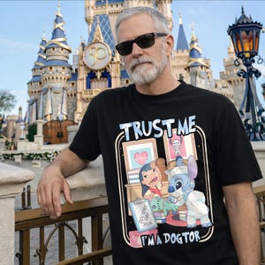 Pu&ograve; includere: T-shirt nera con la scritta "TRUST ME" sopra un'illustrazione a cartoni animati di Lilo e Stitch come dottori. L'illustrazione include il testo "I'M A DOGTOR" e "DOCTORS DAY 2026". Lo sfondo presenta un castello Disney.
