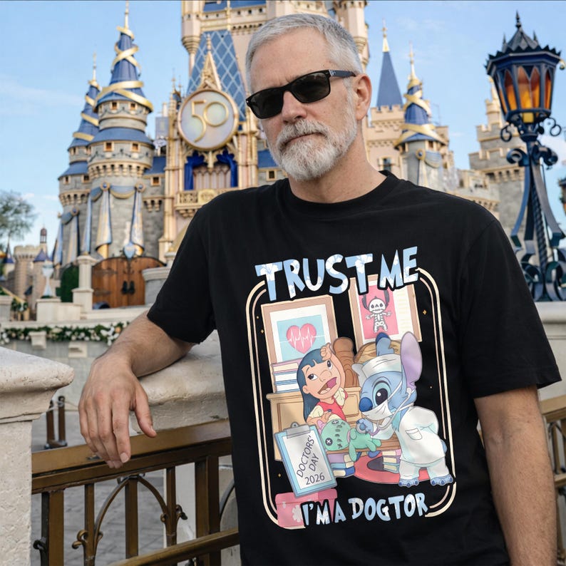 Pu&ograve; includere: T-shirt nera con la scritta "TRUST ME" sopra un'illustrazione a cartoni animati di Lilo e Stitch come dottori. L'illustrazione include il testo "I'M A DOGTOR" e "DOCTORS DAY 2026". Lo sfondo presenta un castello Disney.