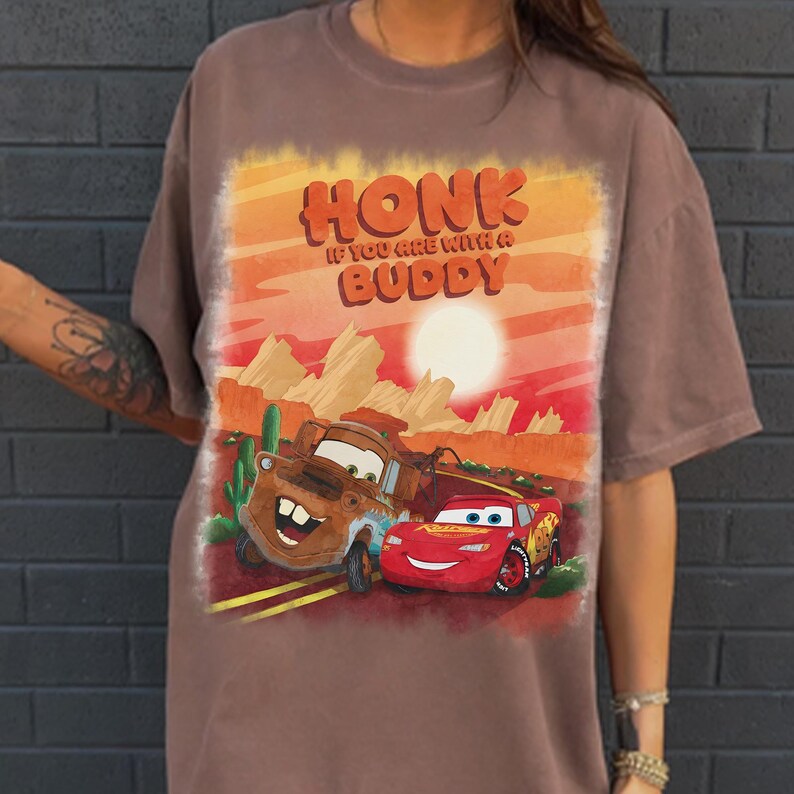 May include: A brown t-shirt featuring a graphic of the characters Mater and Lightning McQueen from the movie Cars, set against a desert landscape with the text "HONK IF YOU ARE WITH A BUDDY".