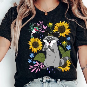 Puede incluir: Una camiseta negra con un mapache de dibujos animados que lleva un sombrero negro con la palabra "Meeko" en él. El mapache está rodeado de girasoles y otras flores.