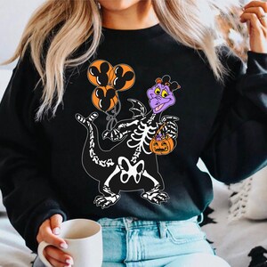 May include: A black long-sleeve sweatshirt featuring a graphic of a purple dragon character wearing a skeleton costume. The dragon holds three orange balloons with black Mickey Mouse silhouettes and an orange jack-o'-lantern candy bucket.