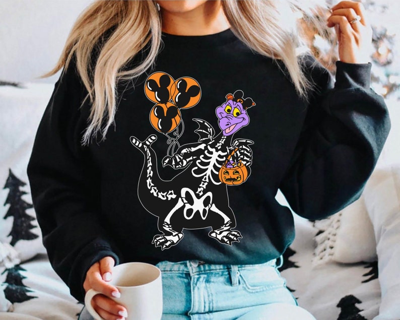 May include: A black long-sleeve sweatshirt featuring a graphic of a purple dragon character wearing a skeleton costume. The dragon holds three orange balloons with black Mickey Mouse silhouettes and an orange jack-o'-lantern candy bucket.