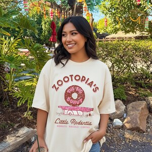 Puede incluir: Camiseta color crema con el texto "ZOOTOPIA'S The Big Donut" sobre un gr&aacute;fico de donut. Debajo, el texto dice "Little Rodentia: Donuts, Bagels, Coffee". La persona sostiene una bebida.