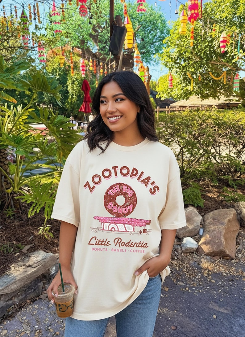 Puede incluir: Camiseta color crema con el texto "ZOOTOPIA'S The Big Donut" sobre un gr&aacute;fico de donut. Debajo, el texto dice "Little Rodentia: Donuts, Bagels, Coffee". La persona sostiene una bebida.