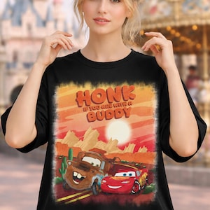 May include: Black t-shirt featuring a graphic of the Cars characters Mater and Lightning McQueen in a desert landscape with the text "HONK IF YOU ARE WITH A BUDDY". The design has a sunset color scheme.