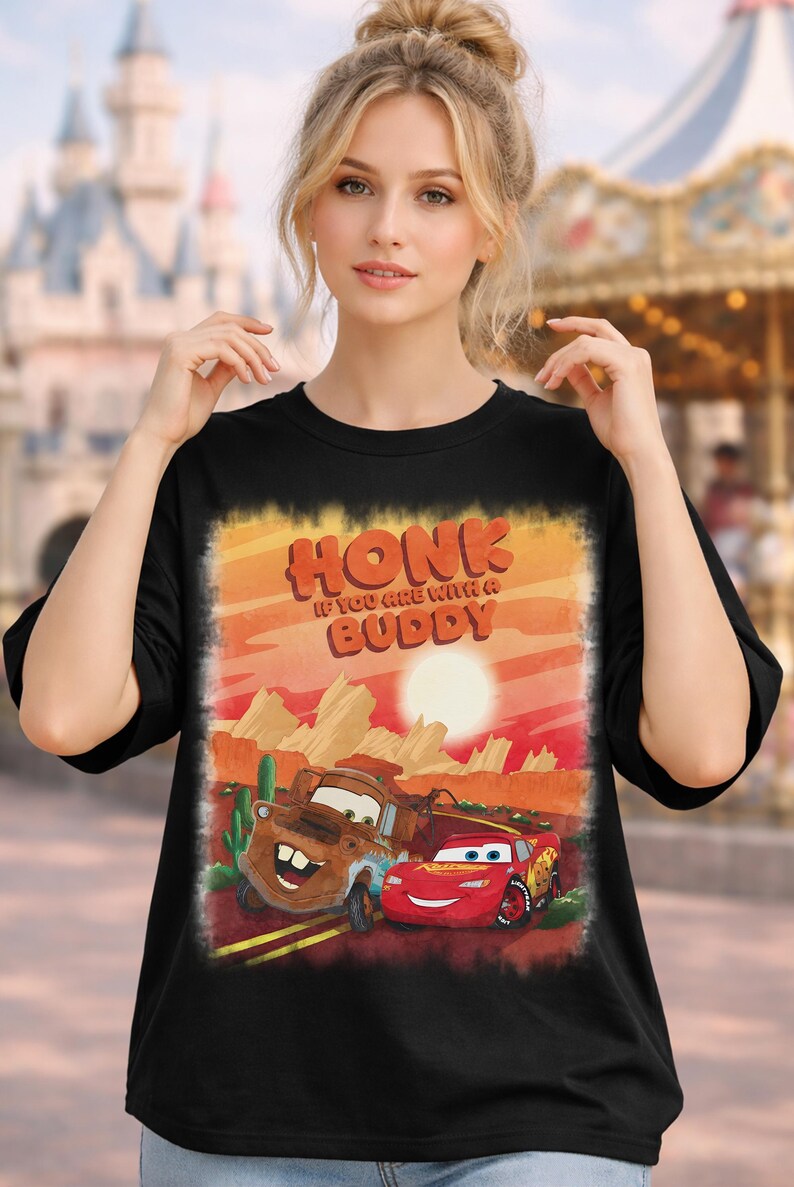May include: Black t-shirt featuring a graphic of the Cars characters Mater and Lightning McQueen in a desert landscape with the text "HONK IF YOU ARE WITH A BUDDY". The design has a sunset color scheme.