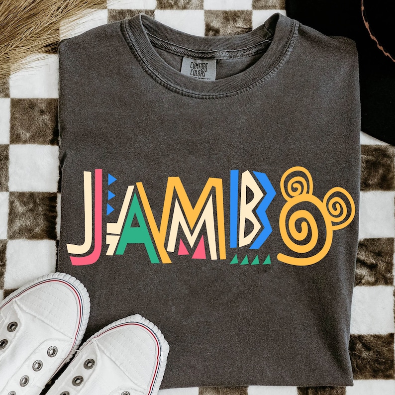Puede incluir: Una camiseta gris oscuro con la palabra "JAMBO" impresa en letras geom&eacute;tricas de colores. Las letras est&aacute;n delineadas en negro y tienen una variedad de formas y patrones. Una oreja de Mickey Mouse en espiral amarilla est&aacute; a la derecha de la palabra.