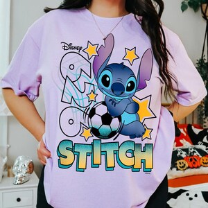 May include: A light purple t-shirt featuring the Disney character Stitch, with the text "GO STITCH" in a colorful design. Stitch is depicted with a soccer ball and yellow stars. The Disney logo is at the top left.