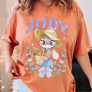 May include: Orange t-shirt with a cartoon illustration of Judy Hopps from Zootopia. The design shows Judy wearing a straw hat and holding a basket of carrots, surrounded by flowers, with the name "JUDY" above.