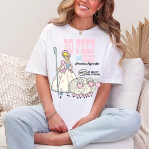 May include: A white t-shirt with a graphic of Bo Peep and her sheep from the Toy Story movie. The text on the shirt reads "Bo Peep and Sheep Porcelain Figure Set Only at Select Toy Stores!"