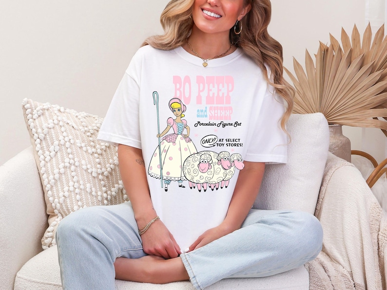 May include: A white t-shirt with a graphic of Bo Peep and her sheep from the Toy Story movie. The text on the shirt reads "Bo Peep and Sheep Porcelain Figure Set Only at Select Toy Stores!"