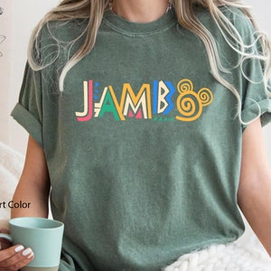 Puede incluir: Una camiseta verde con la palabra "JAMBO" impresa en letras de colores y un dise&ntilde;o de oreja de Mickey Mouse amarillo.