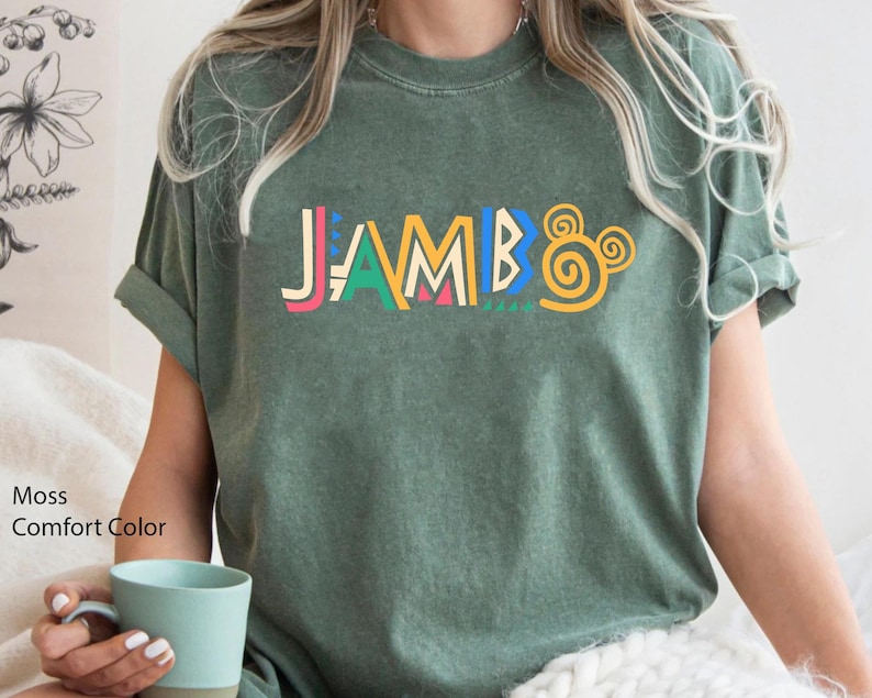 Puede incluir: Una camiseta verde con la palabra "JAMBO" impresa en letras de colores y un dise&ntilde;o de oreja de Mickey Mouse amarillo.