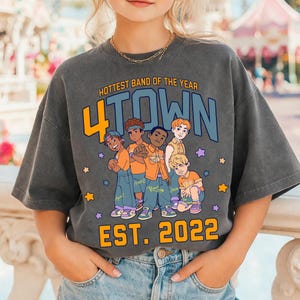 Può includere: T-shirt grigio scuro con la scritta "4TOWN" e "Hottest Band of the Year", più un'illustrazione di cinque ragazzi dei cartoni animati. La maglietta ha anche la scritta "EST. 2022".