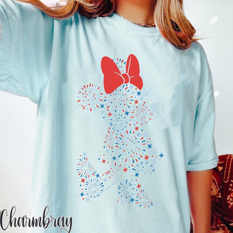 May include: Light blue t-shirt featuring a Minnie Mouse design made of red, white, and blue fireworks. The design includes a large red bow at the top. The word "Charmbray" is printed in cursive at the bottom left.
