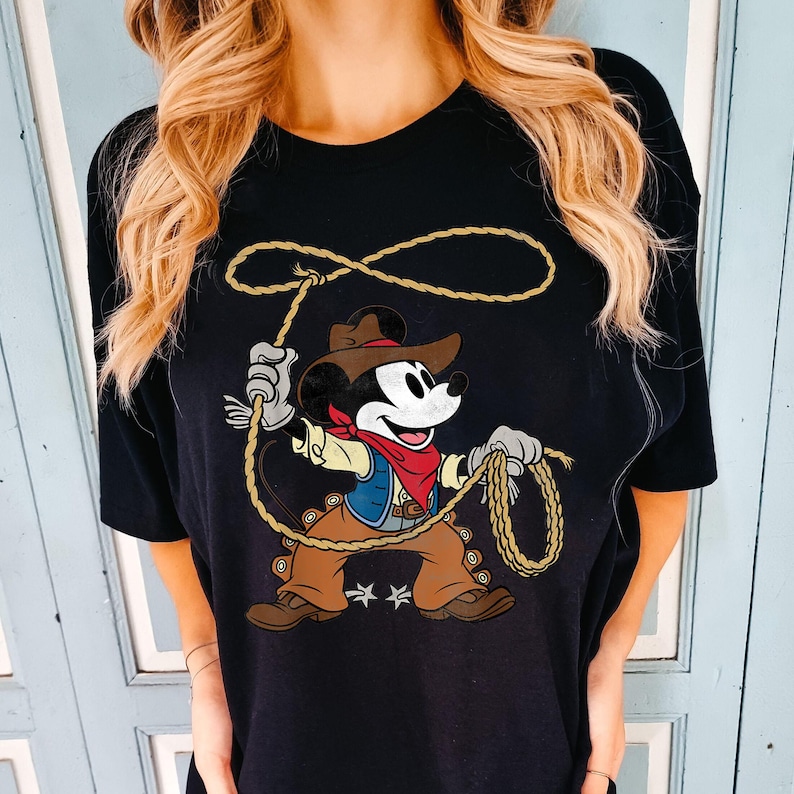 May include: Black t-shirt featuring a cartoon Mickey Mouse dressed as a cowboy, holding a lasso. Mickey wears a brown hat, blue vest, red bandana, and brown pants. The rope is tan, and two white stars are below Mickey.