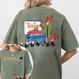Puede incluir: Camiseta verde oliva con una ilustraci&oacute;n de dibujos animados de un zorro vendiendo paletas rosas de un refrigerador azul. El texto "Pavpsicle" est&aacute; encima de las paletas. La camiseta tambi&eacute;n tiene una imagen m&aacute;s peque&ntilde;a del logotipo de Disneyland.