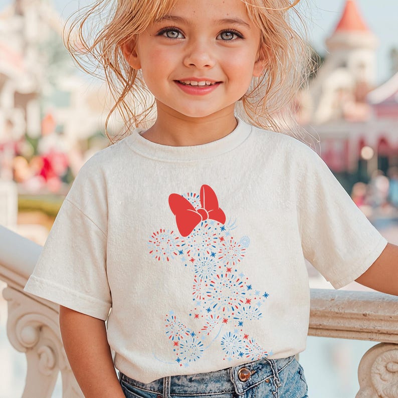 May include: A light beige t-shirt featuring a red bow and a graphic of a character made of red, white, and blue fireworks. The shirt is paired with blue denim jeans. The image is of a young child smiling.