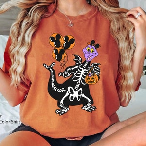 May include: A yam-coloured crewneck sweatshirt featuring a graphic of a purple dragon wearing a skeleton costume. The dragon holds three orange balloons with black Mickey Mouse silhouettes and a small orange jack-o'-lantern candy bucket.