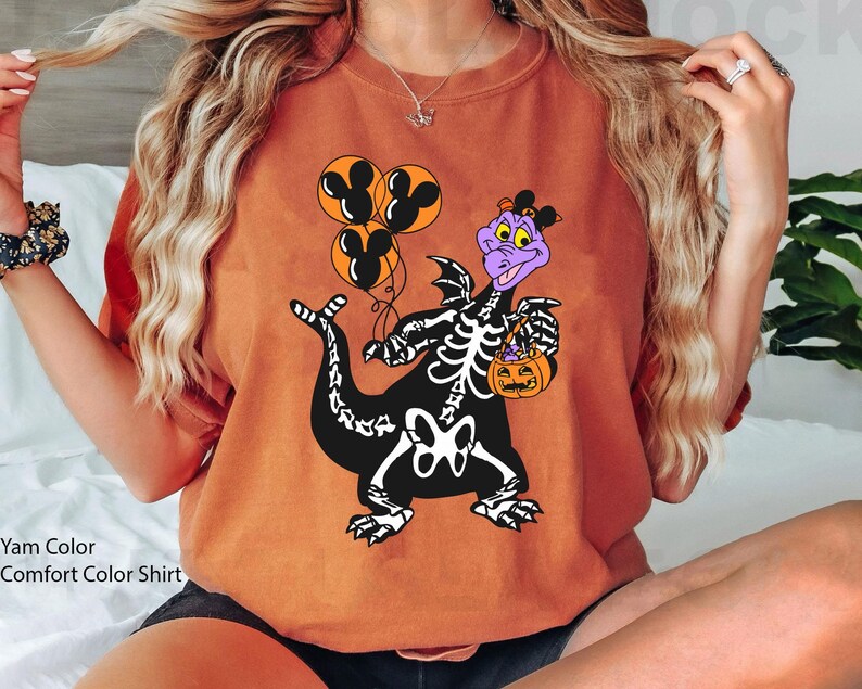 May include: A yam-coloured crewneck sweatshirt featuring a graphic of a purple dragon wearing a skeleton costume. The dragon holds three orange balloons with black Mickey Mouse silhouettes and a small orange jack-o'-lantern candy bucket.