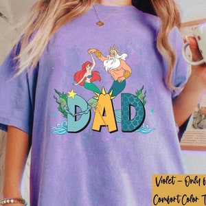 May include: A violet Comfort Color T-shirt with a colourful graphic of Ariel, King Triton, and the word "DAD". The design includes sea elements like seaweed and waves.
