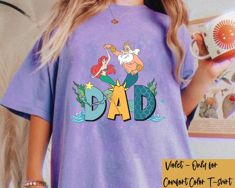 May include: A violet Comfort Color T-shirt with a colourful graphic of Ariel, King Triton, and the word "DAD". The design includes sea elements like seaweed and waves.