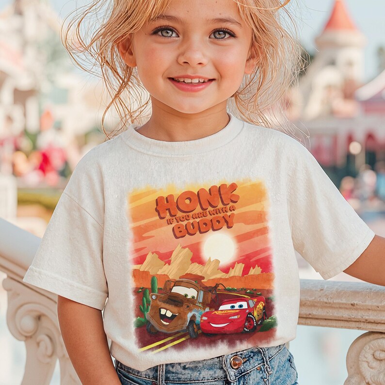 May include: A cream-colored t-shirt featuring a graphic of the characters Mater and Lightning McQueen from the movie Cars, with the text "HONK IF YOU ARE WITH A BUDDY" in orange and red. The design is set against a desert landscape.