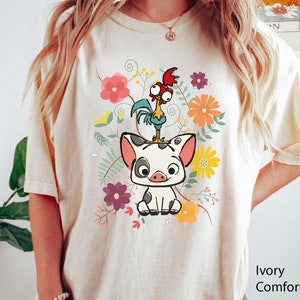 Pu&ograve; includere: Una maglietta bianca con un disegno floreale che presenta un maiale e un gallo dei cartoni animati. Il testo "Ivory Comfort Color" &egrave; visibile nell'angolo in basso a destra dell'immagine.