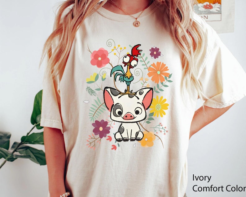 Pu&ograve; includere: Una maglietta bianca con un disegno floreale che presenta un maiale e un gallo dei cartoni animati. Il testo "Ivory Comfort Color" &egrave; visibile nell'angolo in basso a destra dell'immagine.