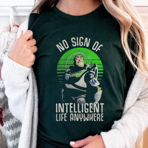 以下が含まれることがあります： トイストーリーの Buzz Lightyear のレトロなグラフィックがプリントされた緑色の T シャツ。グラフィックには「No sign of intelligent life anywhere」というテキストが含まれています。