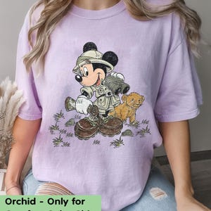 May include: A purple t-shirt with a cartoon image of Mickey Mouse dressed as an explorer with a lion cub. The text "Orchid - Only for Comfort Color Shirt" is at the bottom of the image.