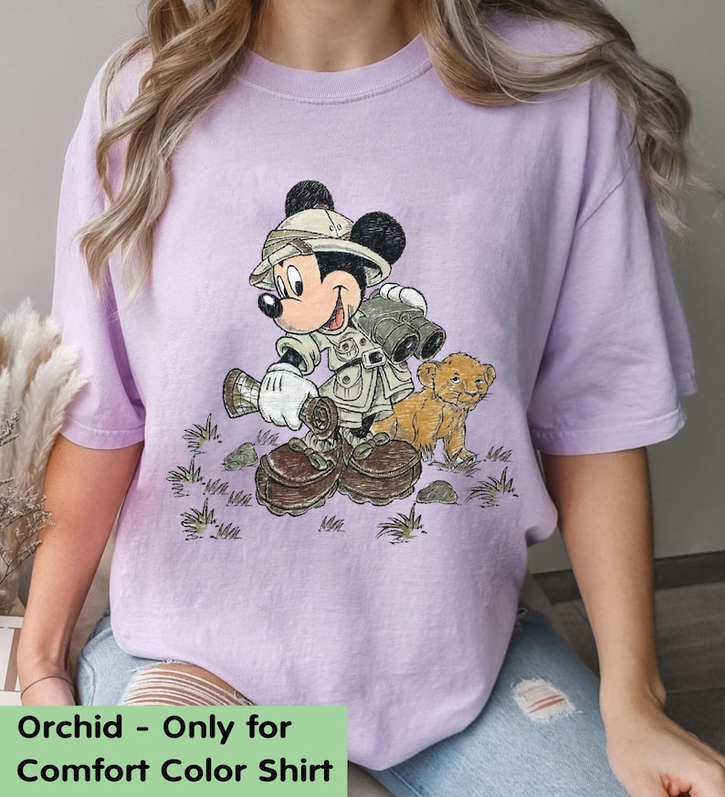 May include: A purple t-shirt with a cartoon image of Mickey Mouse dressed as an explorer with a lion cub. The text "Orchid - Only for Comfort Color Shirt" is at the bottom of the image.
