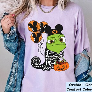 May include: A light purple t-shirt featuring a graphic of a green chameleon wearing a black Mickey Mouse ear hat and a skeleton costume. The chameleon holds three orange Mickey-shaped balloons and an orange jack-o'-lantern bucket filled with sweets.