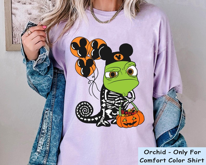 May include: A light purple t-shirt featuring a graphic of a green chameleon wearing a black Mickey Mouse ear hat and a skeleton costume. The chameleon holds three orange Mickey-shaped balloons and an orange jack-o'-lantern bucket filled with sweets.