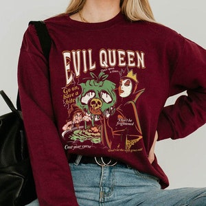 May include: Burgundy sweatshirt with a graphic print of the Evil Queen from Snow White. The graphic features the Evil Queen holding a poisoned apple and the text "Evil Queen, Once upon a time, Go on, have a bite! Don't be frightened, Don't let the witch grow cold, Cast your curse".