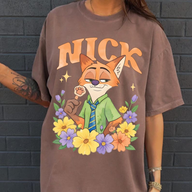 May include: Brown t-shirt with a cartoon fox character named Nick, encircled by colourful flowers. The character wears a green shirt and blue tie, holding a paw-shaped lollipop. The word "NICK" is arched above the fox in orange letters.