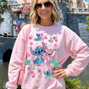May include: Pink sweatshirt featuring a graphic of Stitch and Angel surrounded by flowers. The design includes Stitch in blue and Angel in pink, with floral accents. The sweatshirt is a light pink color.