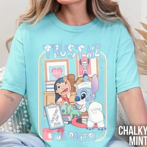 Pu&ograve; includere: T-shirt azzurro con un'illustrazione a cartoni animati. L'illustrazione include una ragazza, un personaggio alieno blu in abito da dottore e il testo "TRUST ME" e "I'M A DOCTOR". &Egrave; visibile anche la scritta "DOCTORS DAY 2026".
