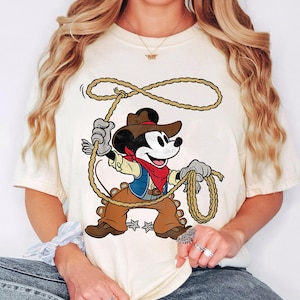 May include: Cream-colored t-shirt featuring a cartoon Mickey Mouse dressed as a cowboy, holding a lasso. Mickey wears a brown hat, vest, and pants, with a red bandana. The design is centered on the shirt, showcasing a playful, Western theme.