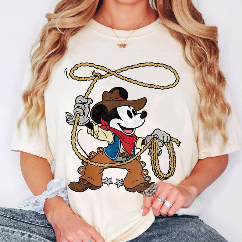 May include: Cream-colored t-shirt featuring a cartoon Mickey Mouse dressed as a cowboy, holding a lasso. Mickey wears a brown hat, vest, and pants, with a red bandana. The design is centered on the shirt, showcasing a playful, Western theme.