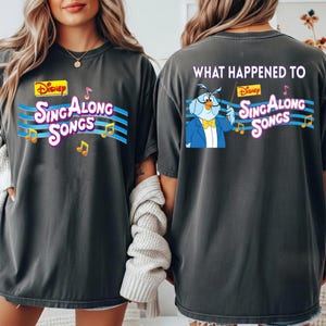 May include: Dark gray t-shirt featuring the Disney "Sing Along Songs" logo on the front and back. The front design includes the logo with musical notes. The back features an owl character and the text "What Happened To".