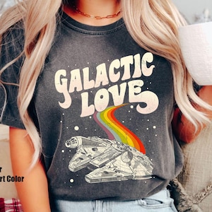 Puede incluir: Camiseta gris oscuro con la frase "GALACTIC LOVE" en una fuente retro. Un arcoíris conecta el texto con una ilustración detallada de una nave espacial. La camiseta está etiquetada como "Pepper Comfort Color".