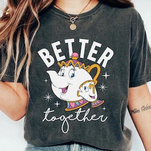 May include: Dark gray t-shirt featuring a cartoon teapot and teacup with the text "Better together" in white script. The teapot and teacup are white with gold, pink, and blue accents. The shirt has a vintage, worn look.